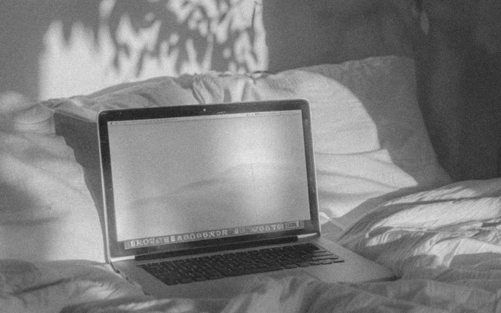 Black and white grainy image of a Mac laptop sitting on a white, messy bed. The laptop relates to answering the question "what is website copywriting"? 